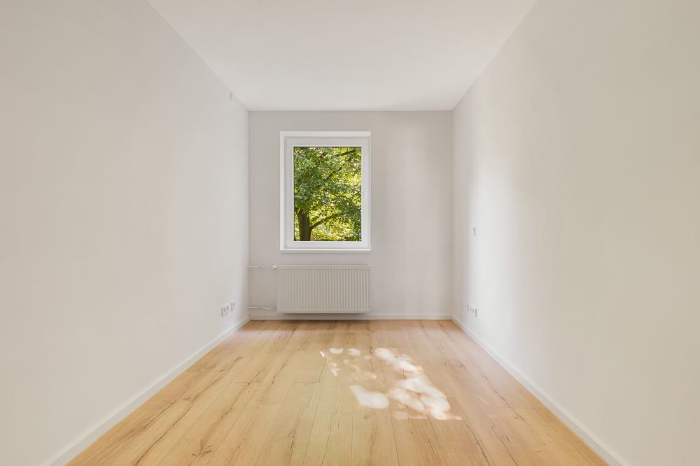 Heller, leerer Raum mit Holzfußboden, weißem Fenster und Blick auf Bäume.