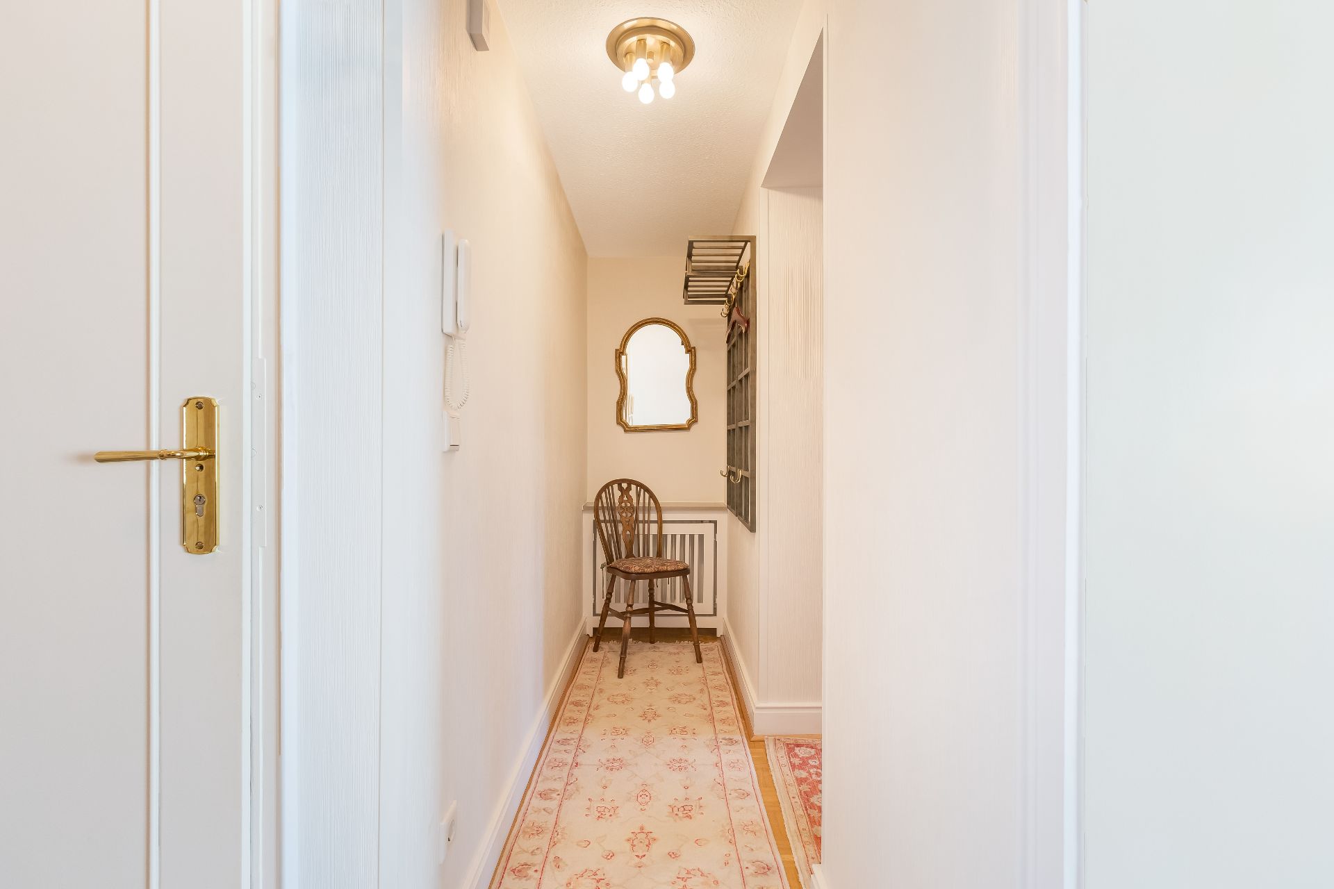 Schmaler Flur mit Teppich, Holzstuhl, Spiegel und Garderobe an der Wand.