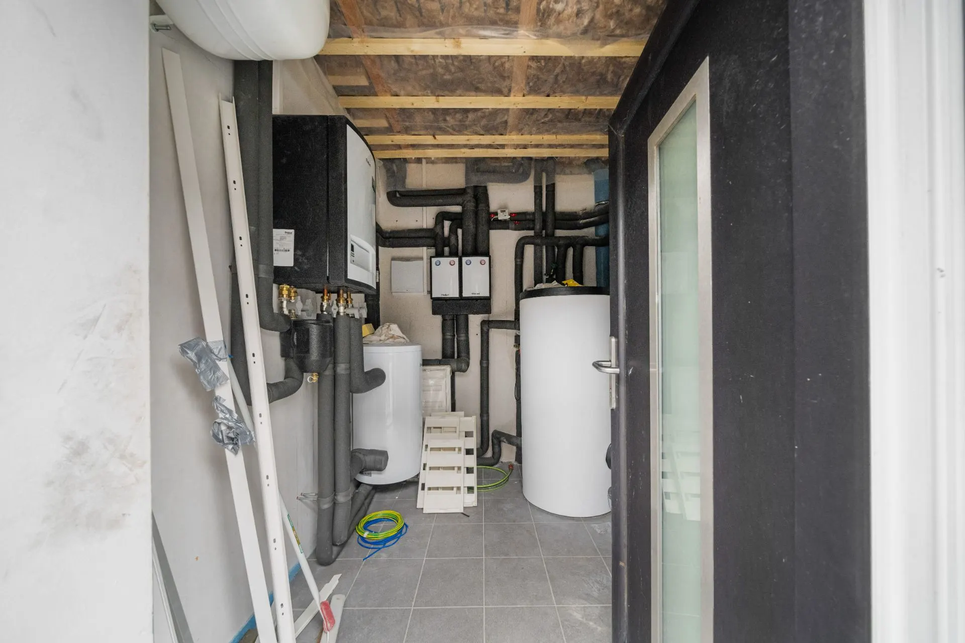 Hauswirtschaftsraum mit Heizungsanlage, Rohren und weißen Kesseln auf grauem Fliesenboden.