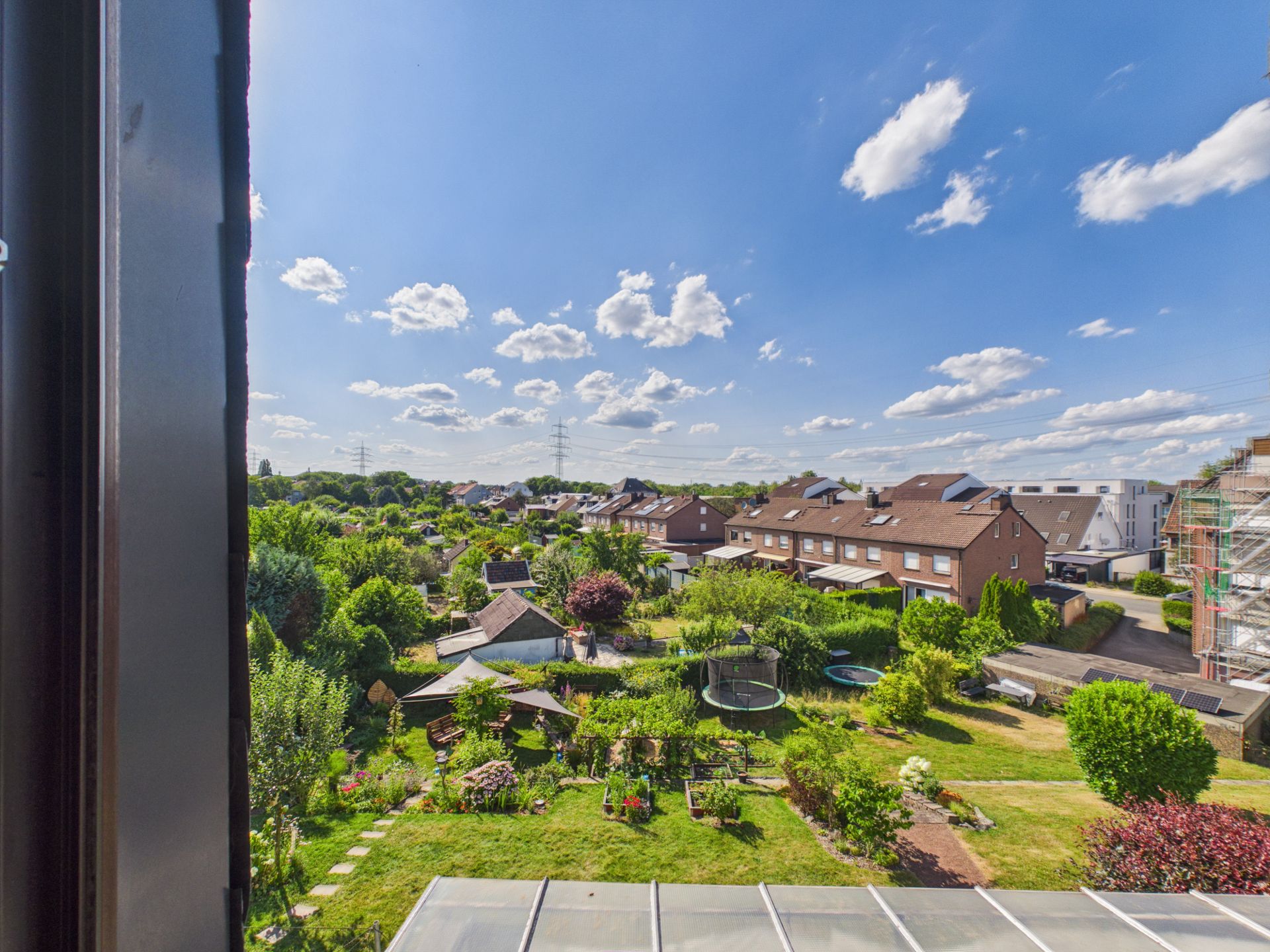 Blick aus dem Fenster auf einen grünen Garten mit Wohnhäusern im Hintergrund unter blauem Himmel.