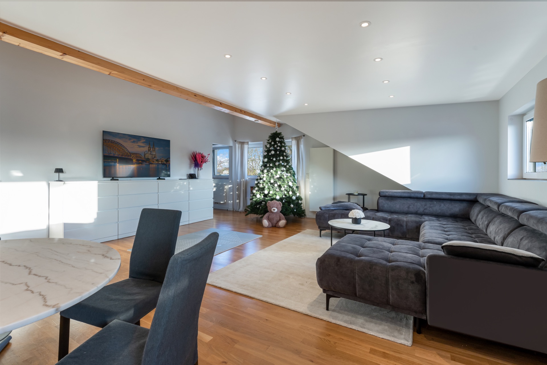 Helles Wohnzimmer mit großer Eckcouch, rundem Tisch und geschmücktem Weihnachtsbaum.