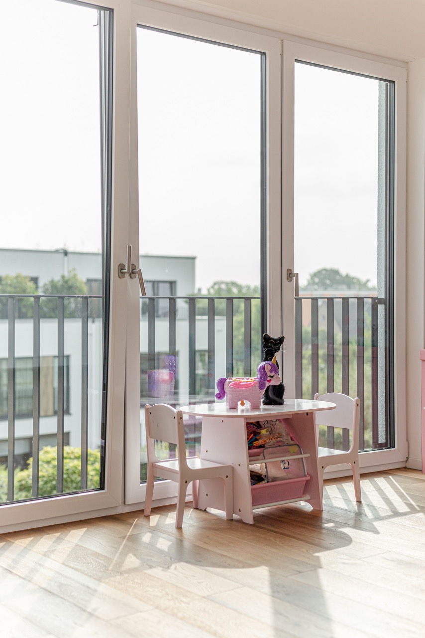 Kleiner Kindertisch mit zwei Stühlen und Spielzeug vor bodentiefen Fenstern mit Blick nach draußen.