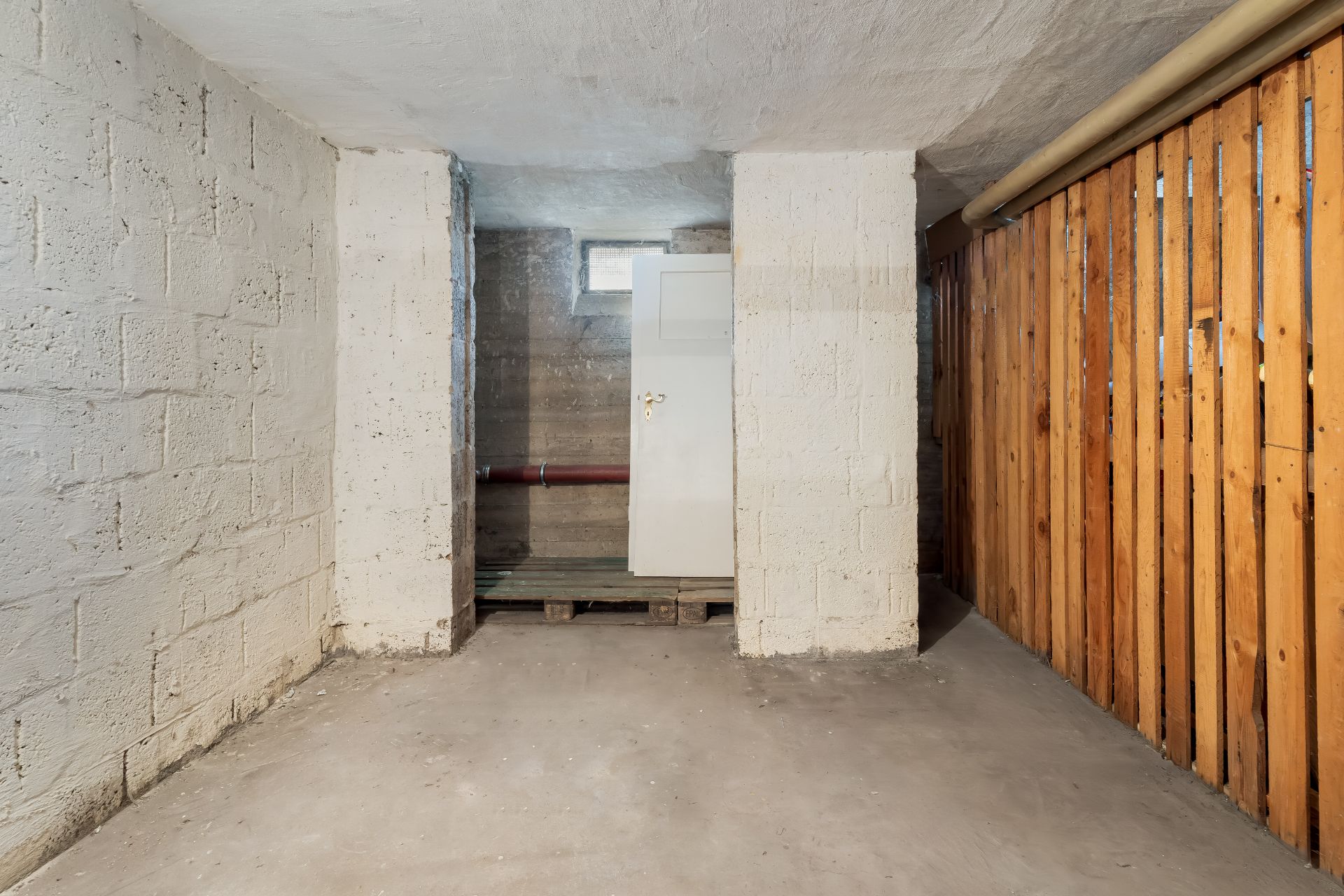 Leerer Kellerraum mit Betonwänden, Tür, kleinem Fenster und Holzwand.