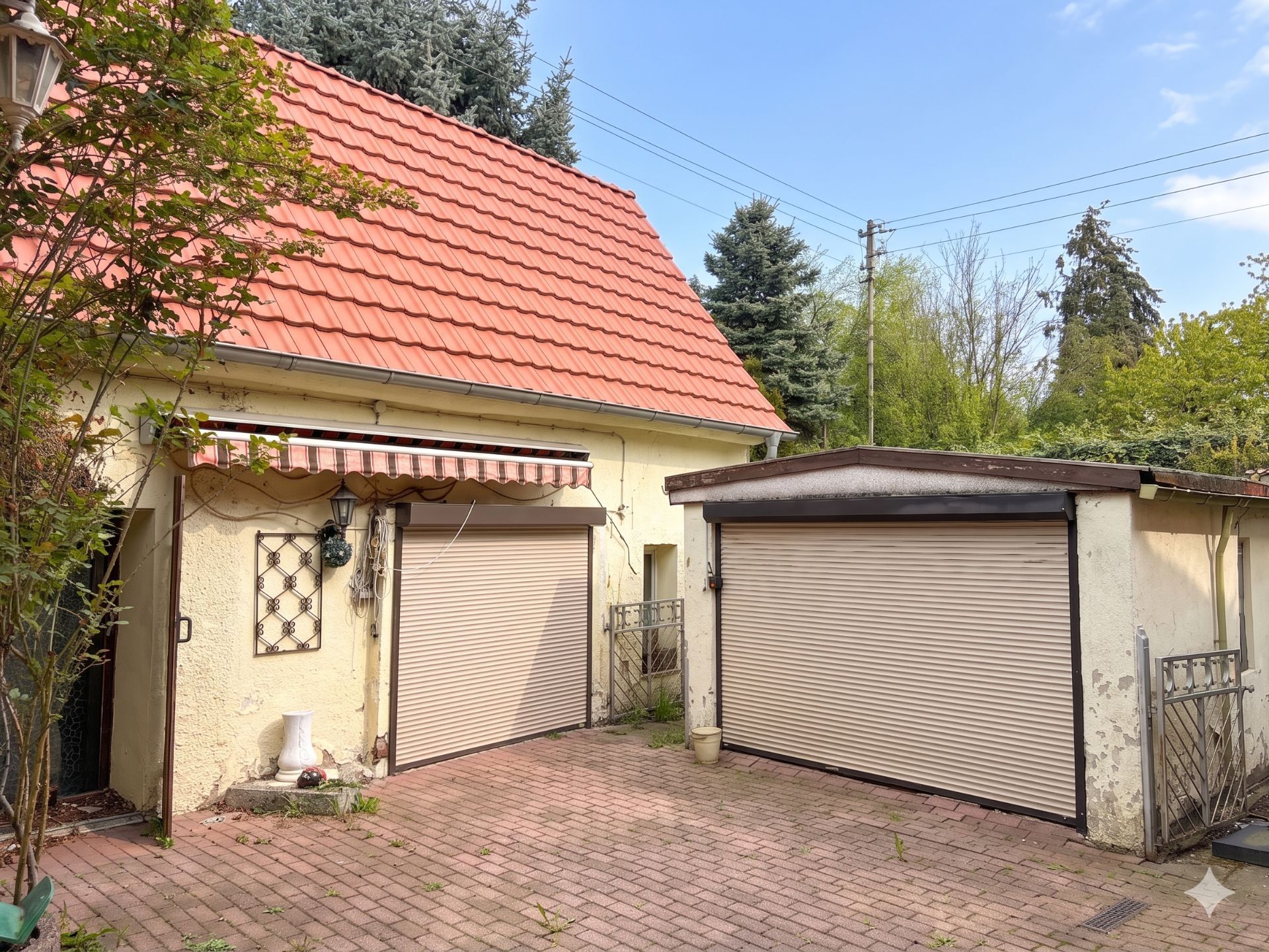 Zwei Garagen mit geschlossenen Toren vor einem Haus mit rotem Dach und gepflastertem Hof.