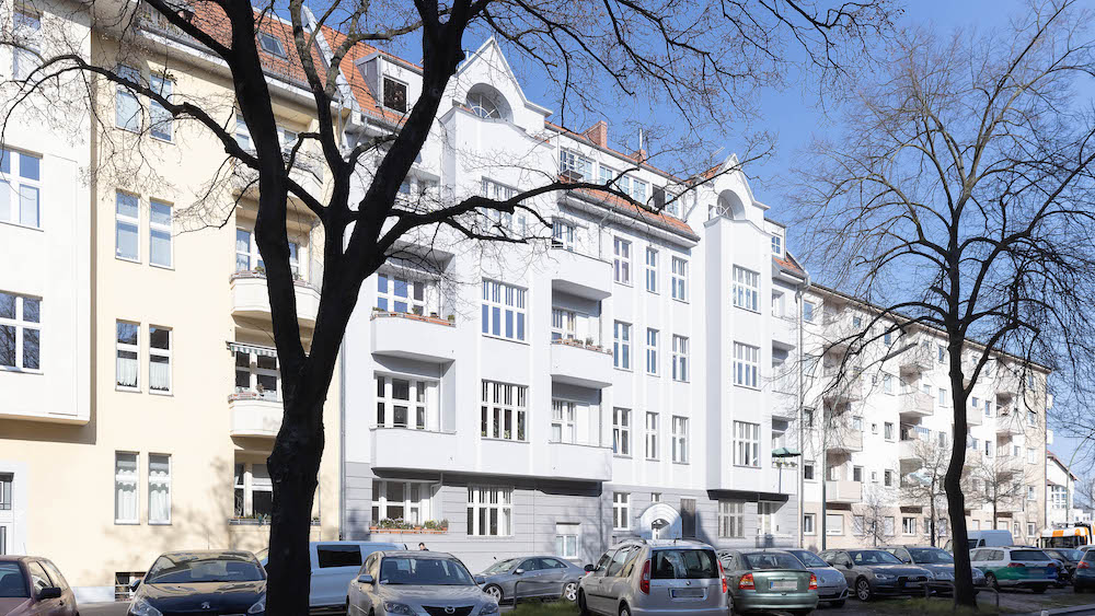 Wohnhaus mit mehreren Stockwerken, Balkonen und parkenden Autos davor, Bäume im Vordergrund.