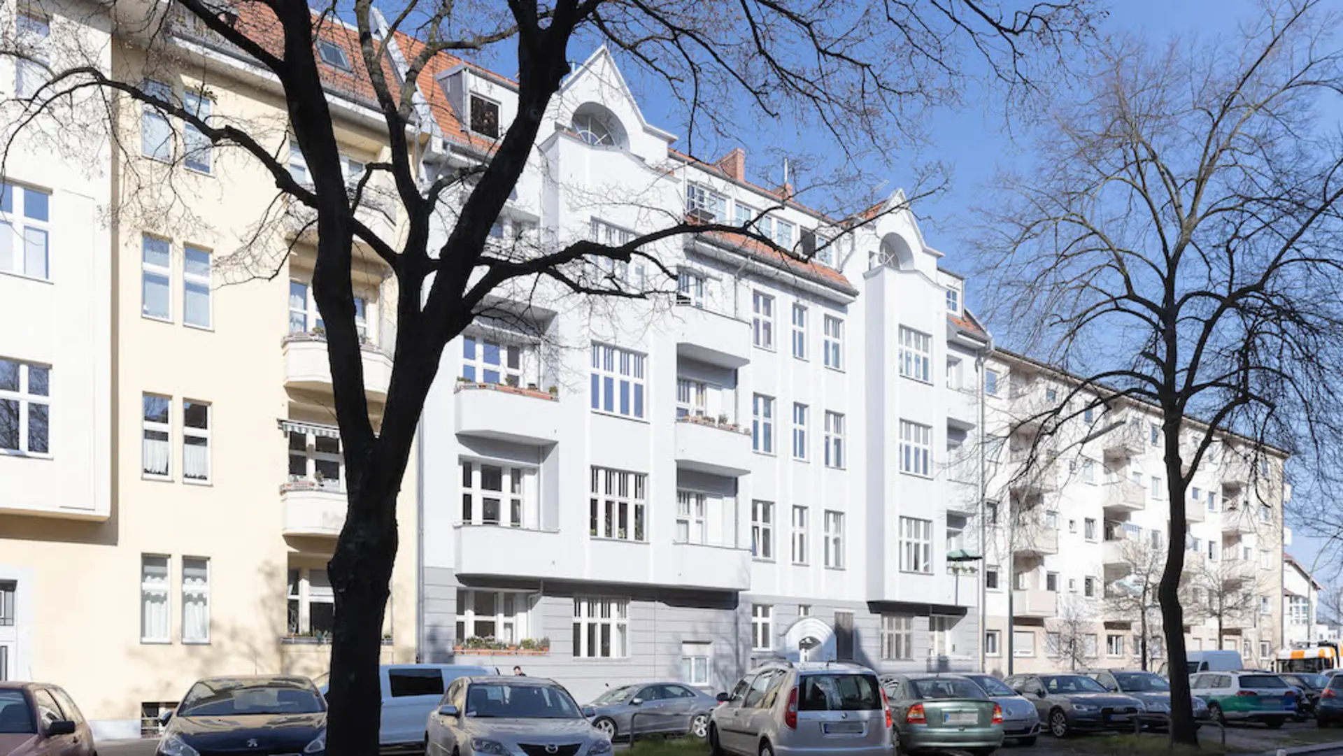 Wohnhaus mit mehreren Stockwerken, Balkonen und parkenden Autos davor, Bäume im Vordergrund.