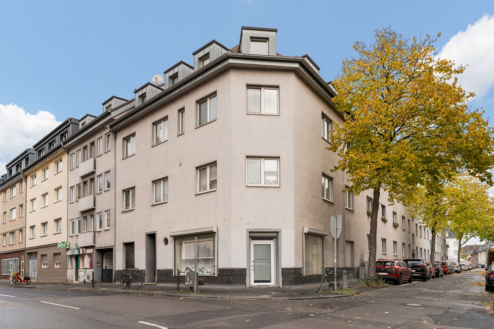 Eckhaus mit mehreren Stockwerken, Fenstern und eingeschossigem Dach, an einer Straßenecke mit Bäumen.