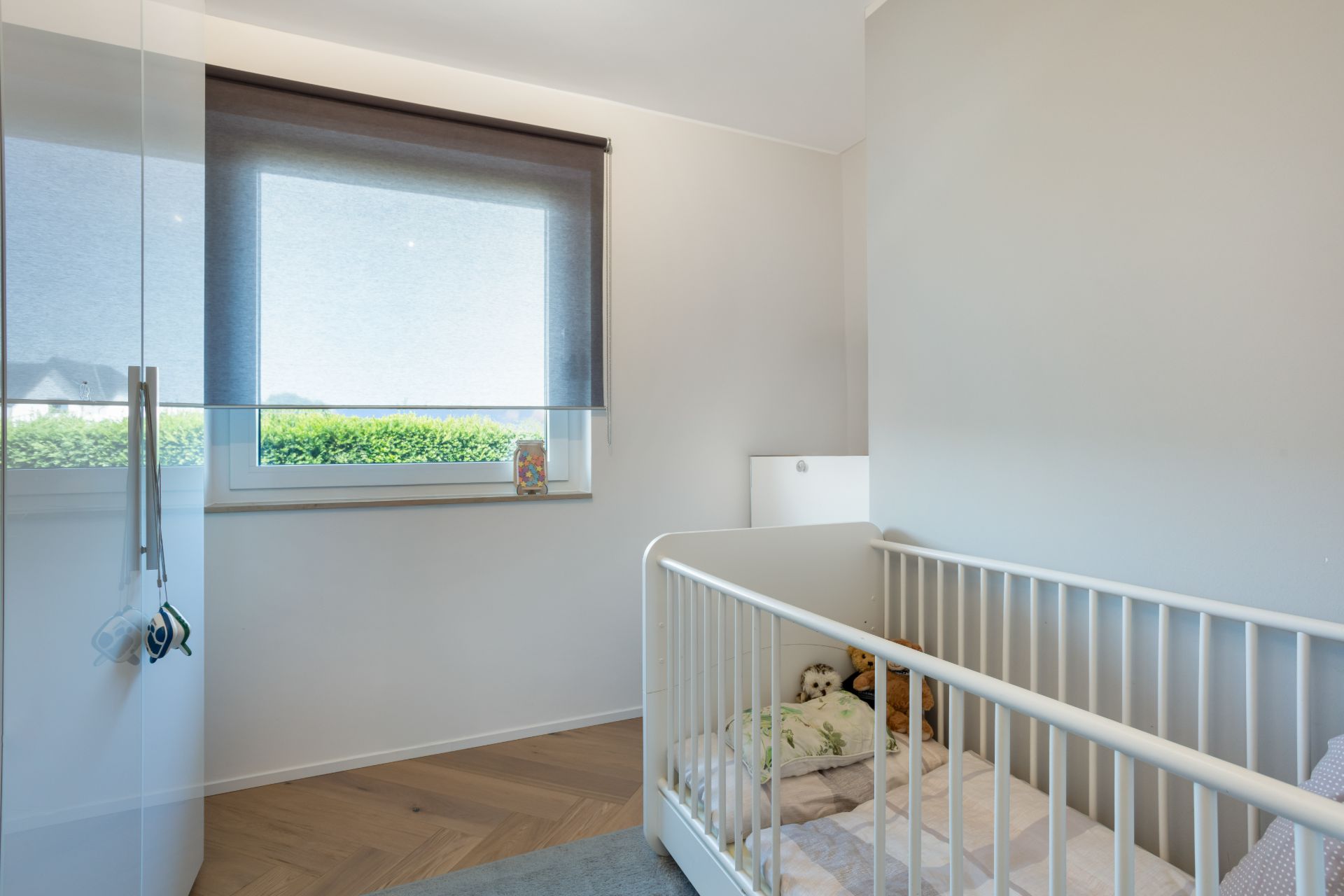 Kleines Kinderzimmer mit Gitterbett, Spielzeug und Fenster mit halb geschlossenem Rollo.