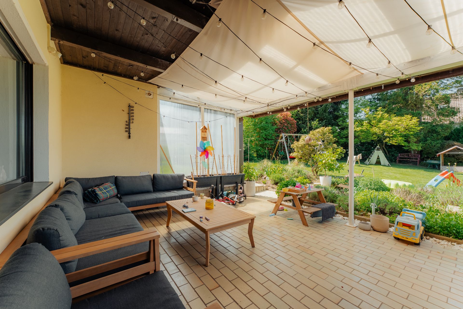 Überdachte Terrasse mit Sitzgruppe, kleinem Tisch, Kinderspielzeug und Blick in den Garten.