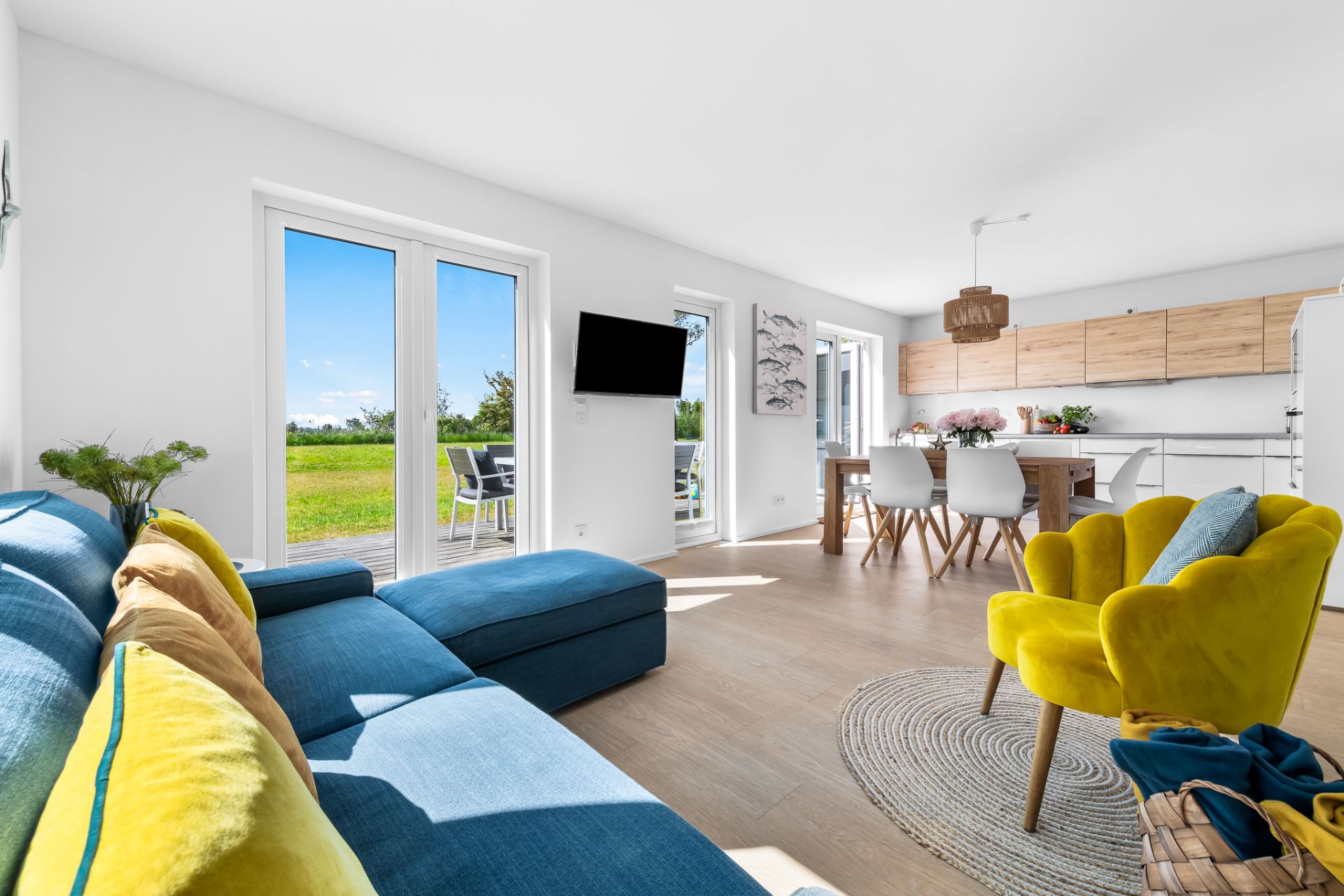 Helles Wohnzimmer mit blauer Couch, gelbem Sessel, Essbereich und Zugang zur Terrasse mit Blick ins Grüne.