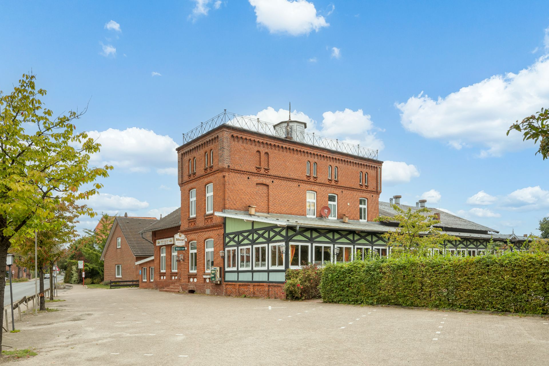 Backsteinwohnhaus mit Wintergarten und Parkplatz, umgeben von Bäumen, unter blauem Himmel.