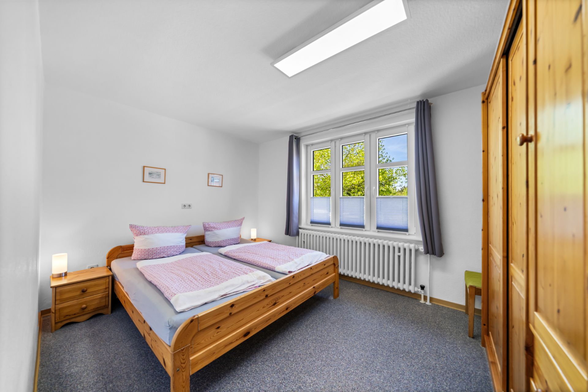 Helles Schlafzimmer mit Doppelbett, Holzschrank und großem Fenster mit grauen Vorhängen.