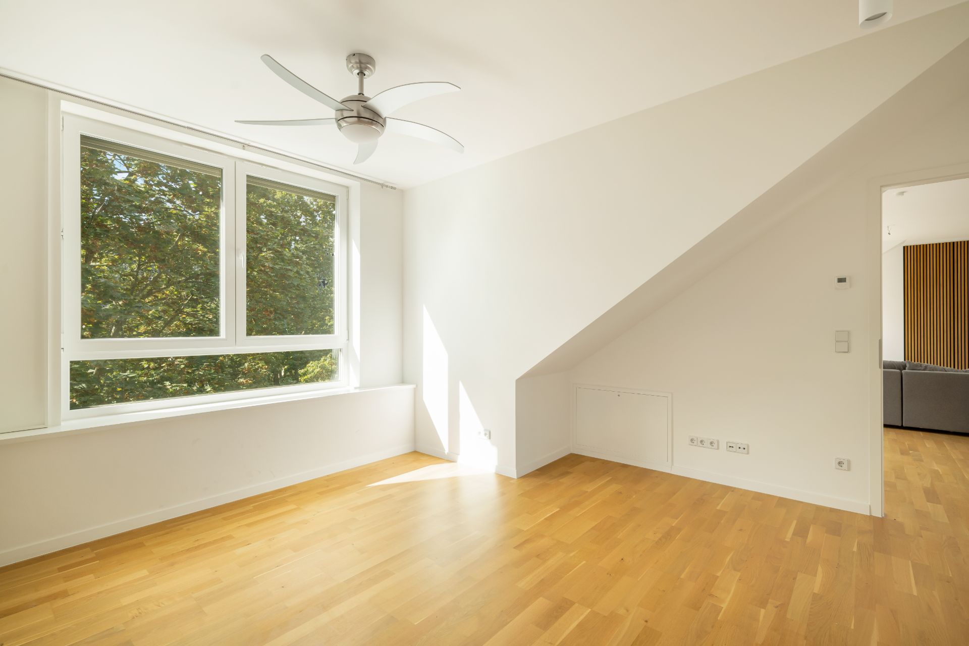 Helles Zimmer mit Holzfußboden, großem Fenster und Deckenventilator, Tür zu weiterem Raum sichtbar.