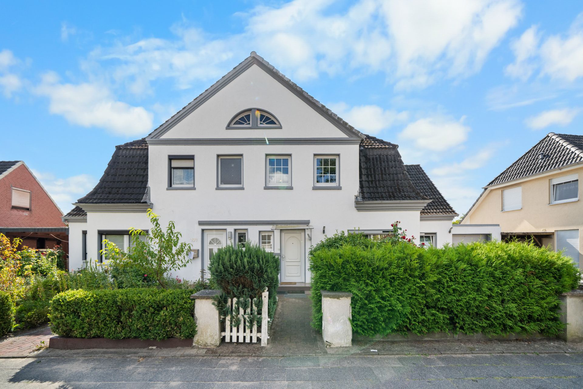 Doppelhaus mit weißer Fassade, dunklem Dach und kleinem Vorgarten mit Hecke und Zaun.