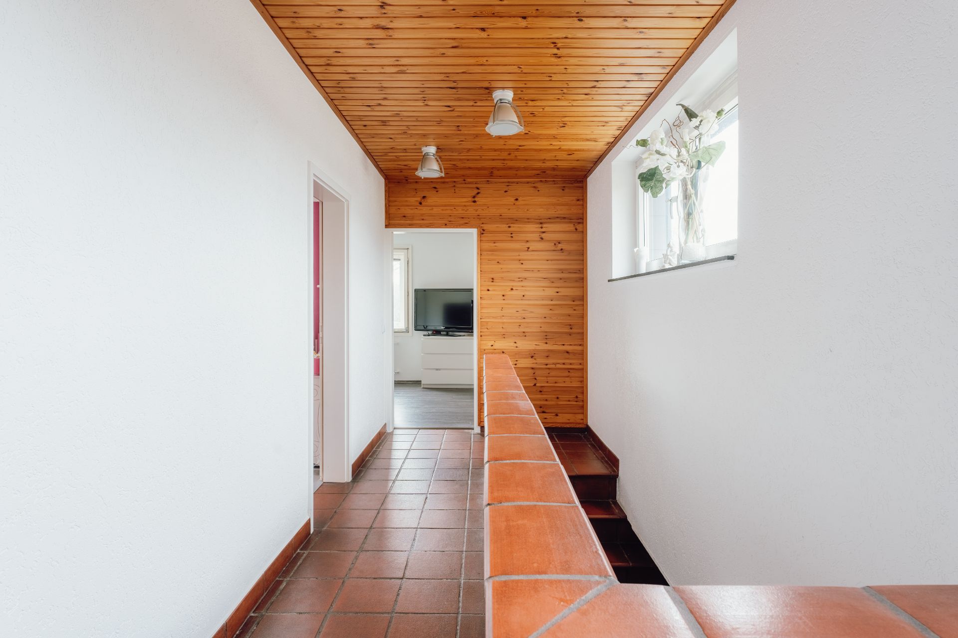 Heller Flur mit Terrakottafliesen, Holzdecke und Fenster mit Blumenvase.