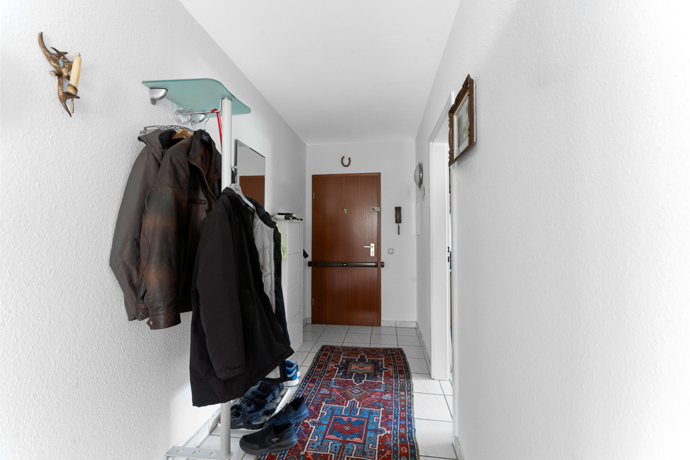 Flur mit Kleiderständer, mehreren Jacken, Schuhen, Teppich und brauner Haustür.