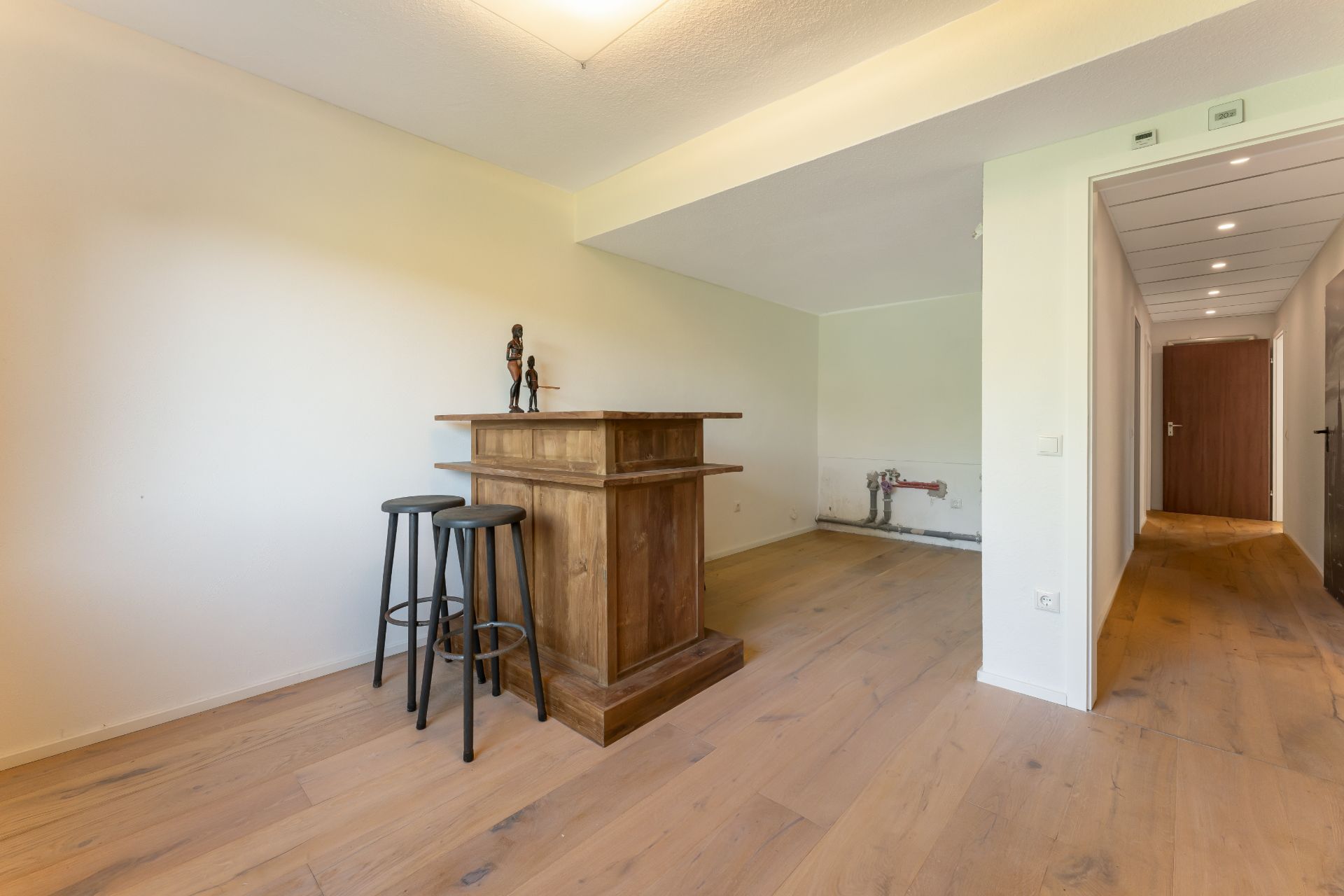 Heller Raum mit Holzboden, kleiner Holztheke und zwei Hockern, neben einem Flur.