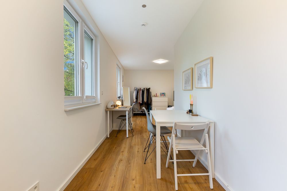 Heller Raum mit Holzfußboden, zwei Schreibtischen, Stühlen und Fenster, im Hintergrund Kommode und Garderobe.