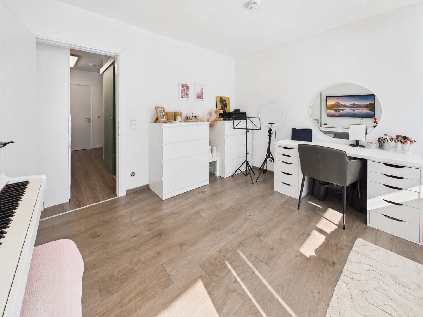 Helles Zimmer mit Schminktisch, Kommode, Klavier, Stuhl und Musikständer auf Holzfußboden.