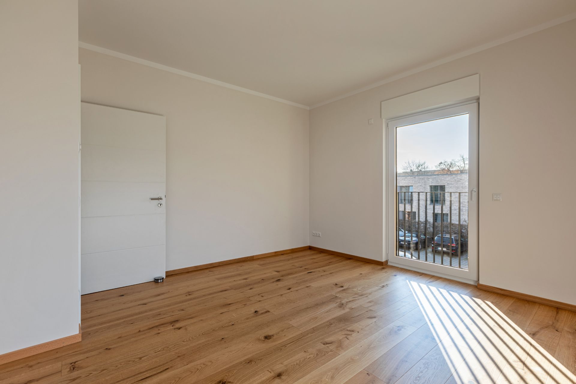 Leeres Zimmer mit Holzboden, weißer Tür und bodentiefem Fenster mit Gitter.