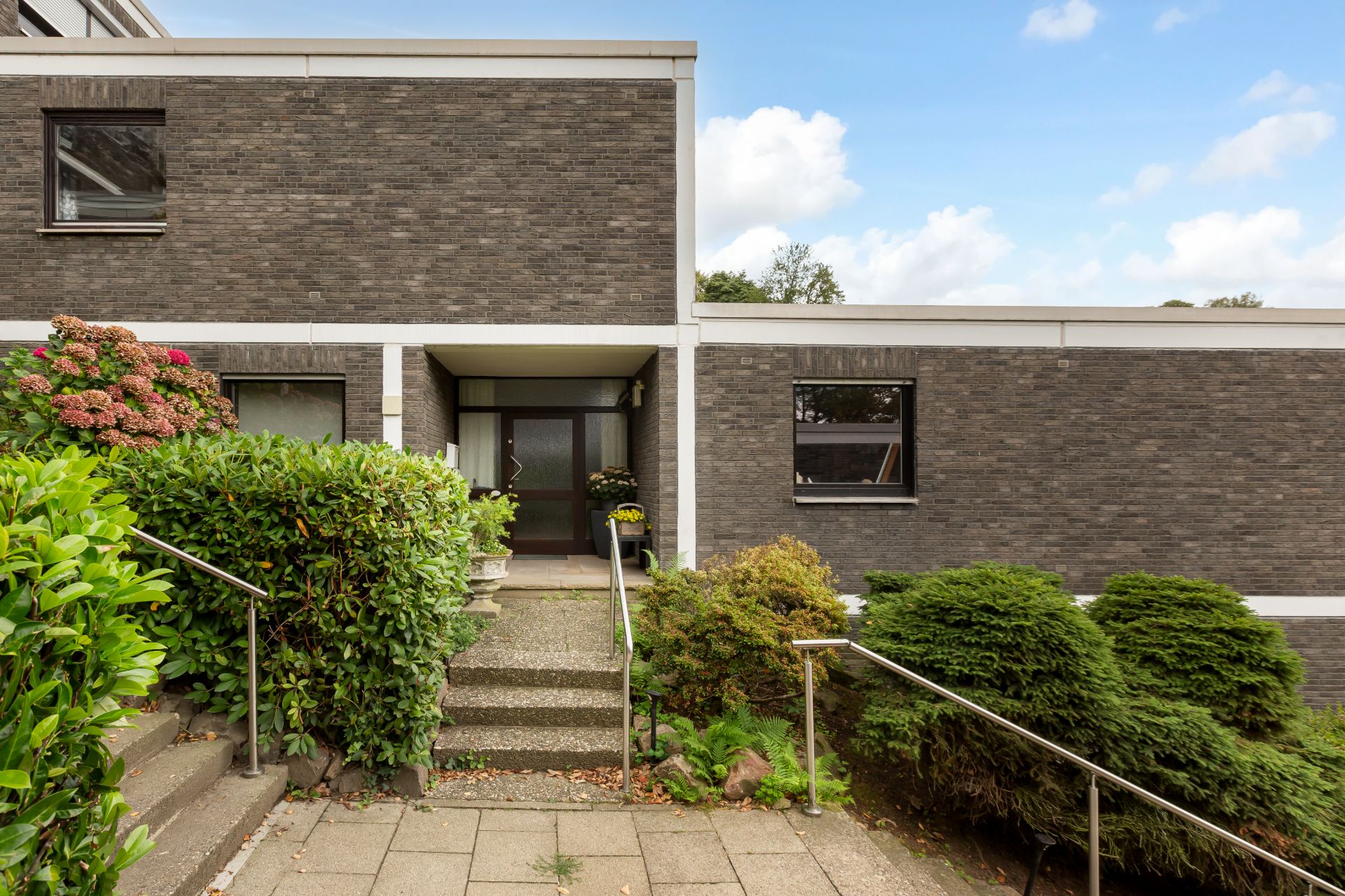 Eingang eines modernen Wohnhauses mit grauer Klinkerfassade, Treppe und Pflanzen.