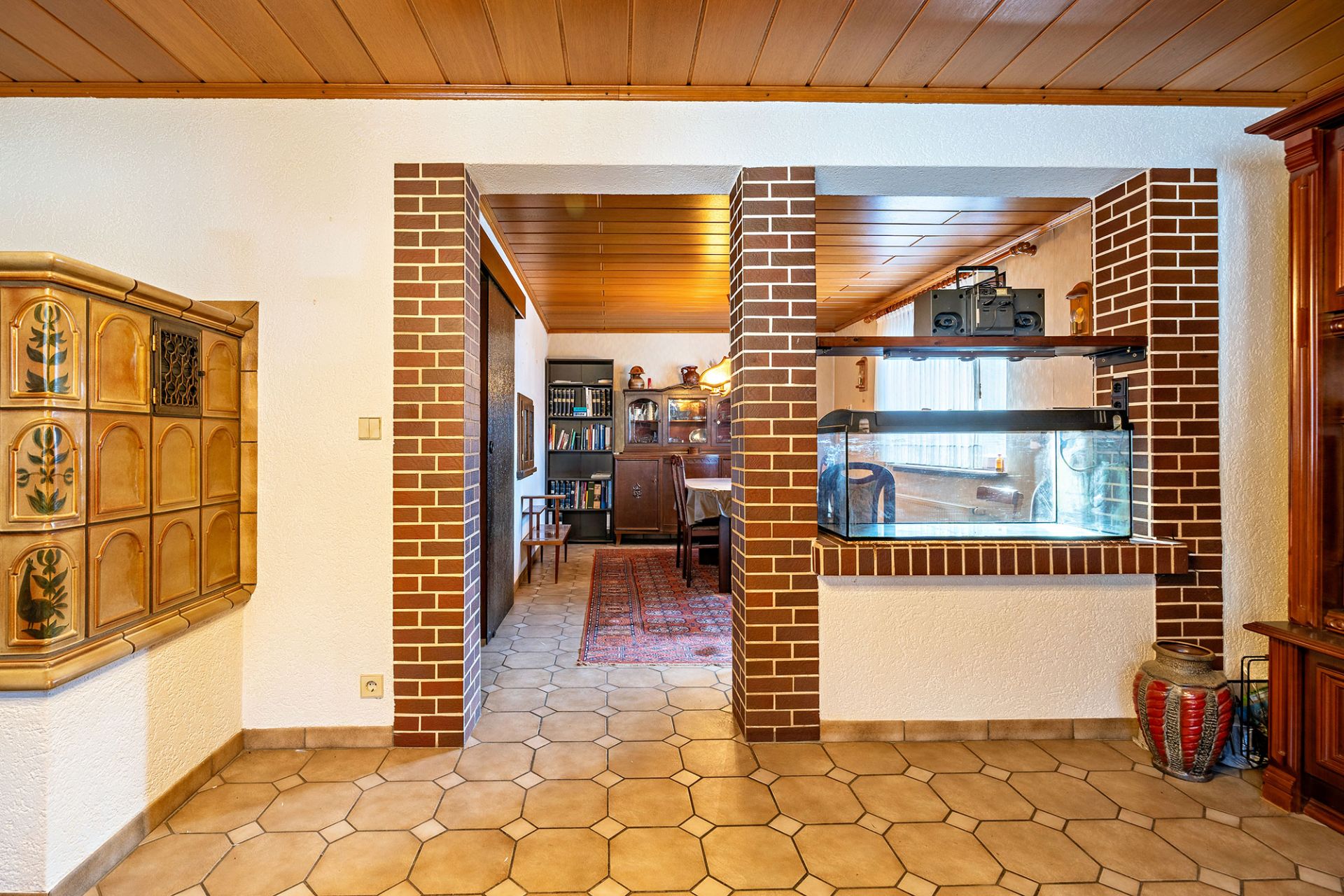 Wohnzimmer mit Kachelofen, Aquarium, Holzdecke und Durchgang zu Esszimmer mit Regal und Teppich.