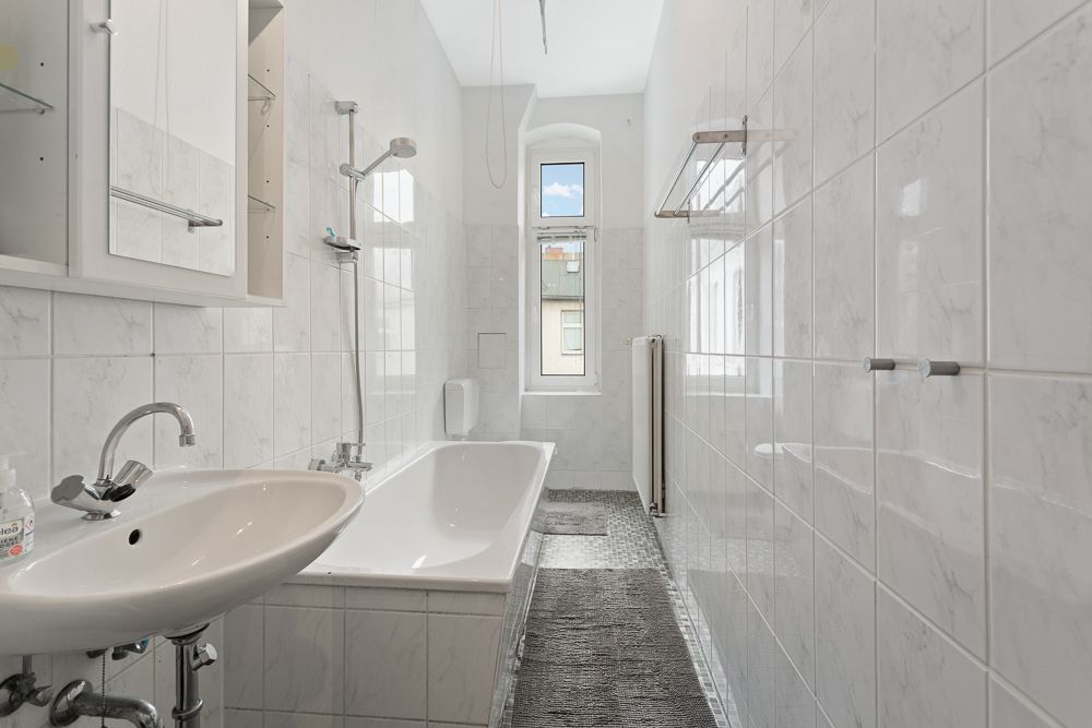 Helles Badezimmer mit Waschbecken, Badewanne, Dusche, Fenster und weißen Fliesen.