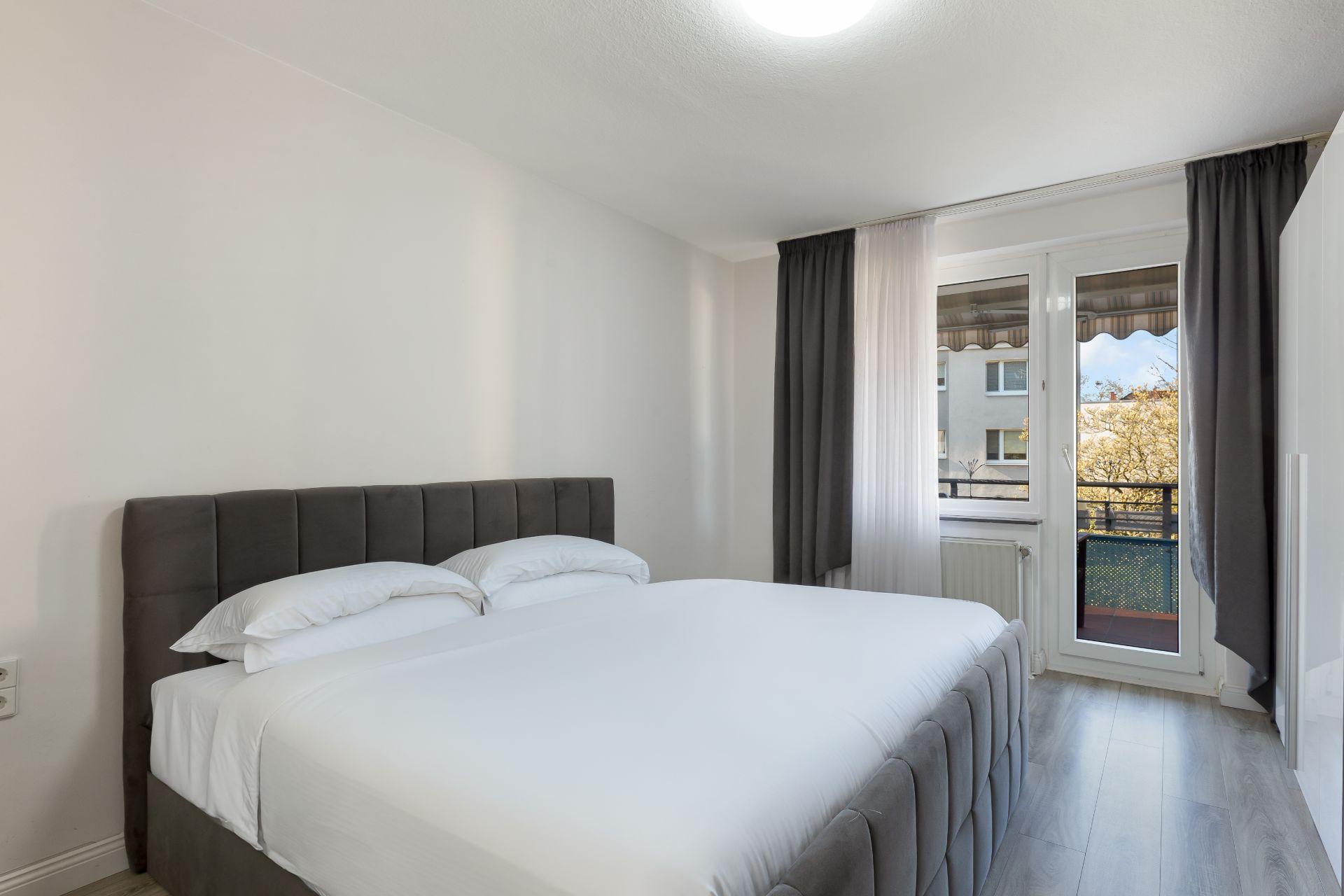 Schlafzimmer mit Doppelbett, Fenstertür und Vorhängen, heller Boden und neutraler Wandfarbe.