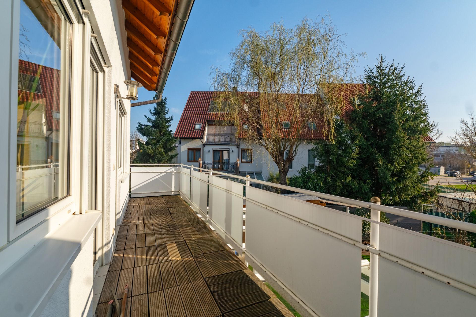 Balkon mit Holzboden und weißem Geländer, Blick auf Nachbarhäuser und Bäume.