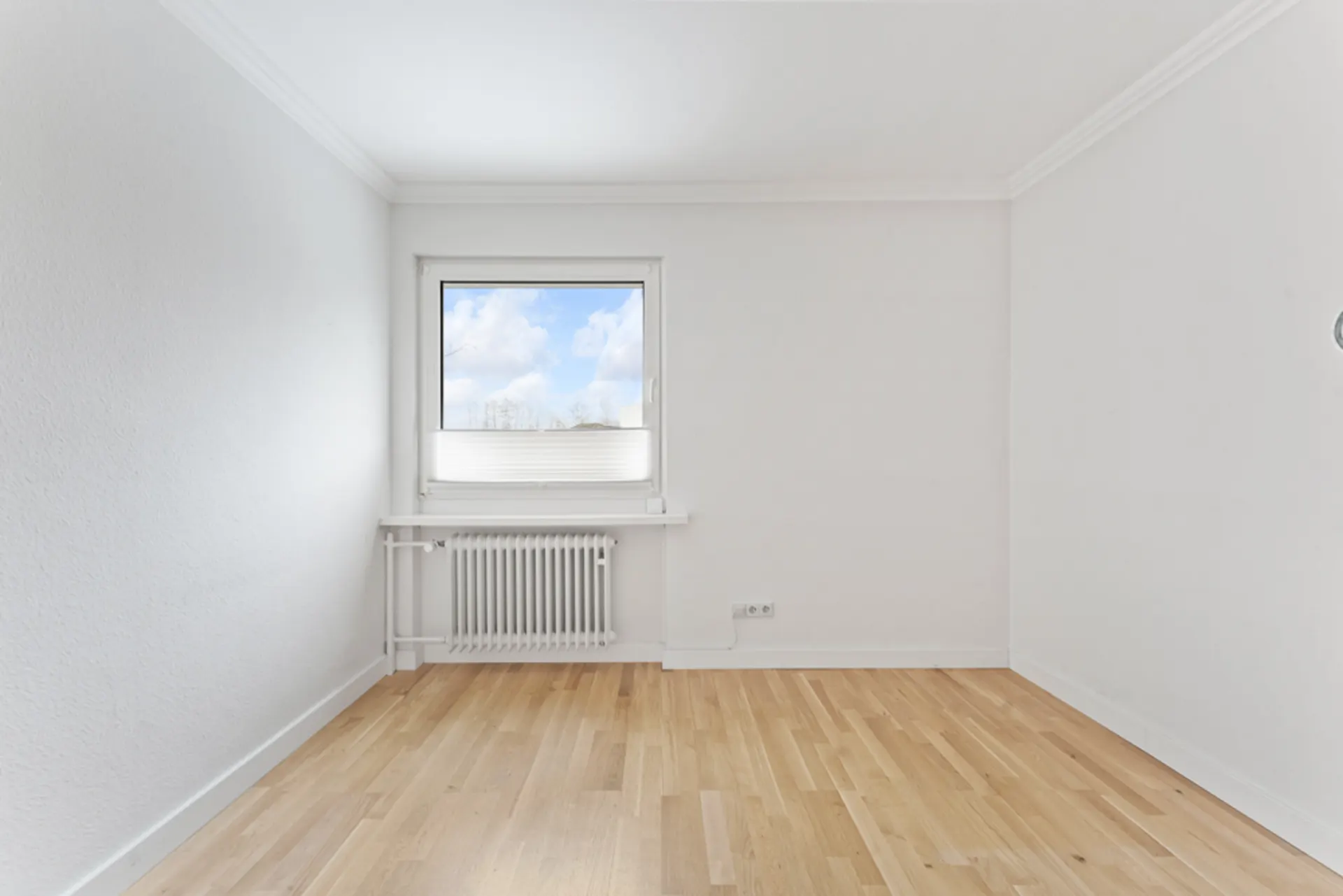 Leerer, heller Raum mit Holzfußboden, weißer Wand, Fenster und Heizkörper.