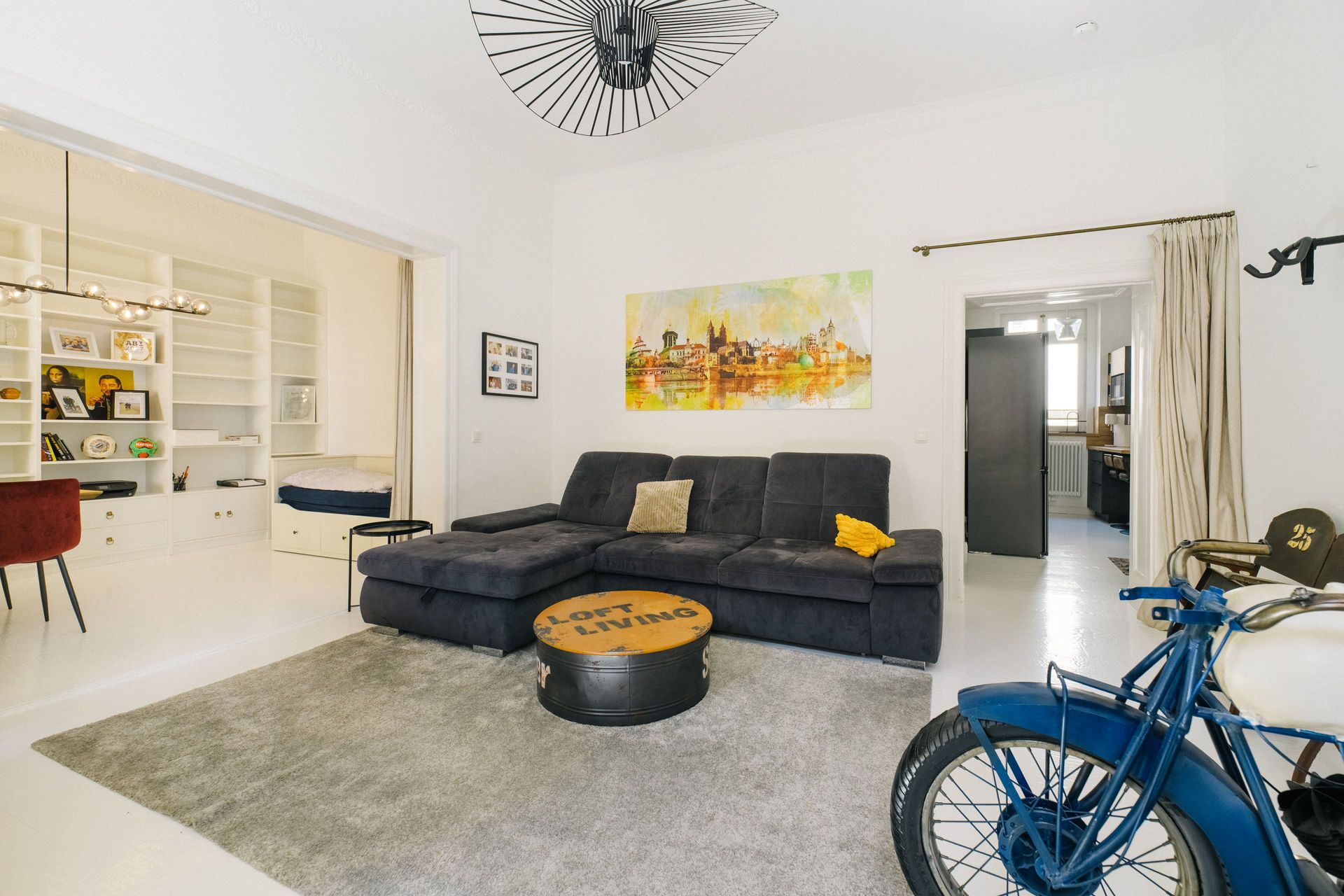 Helles Wohnzimmer mit dunkelgrauem Sofa, Teppich, Bild an der Wand und einem Motorrad in der Ecke.