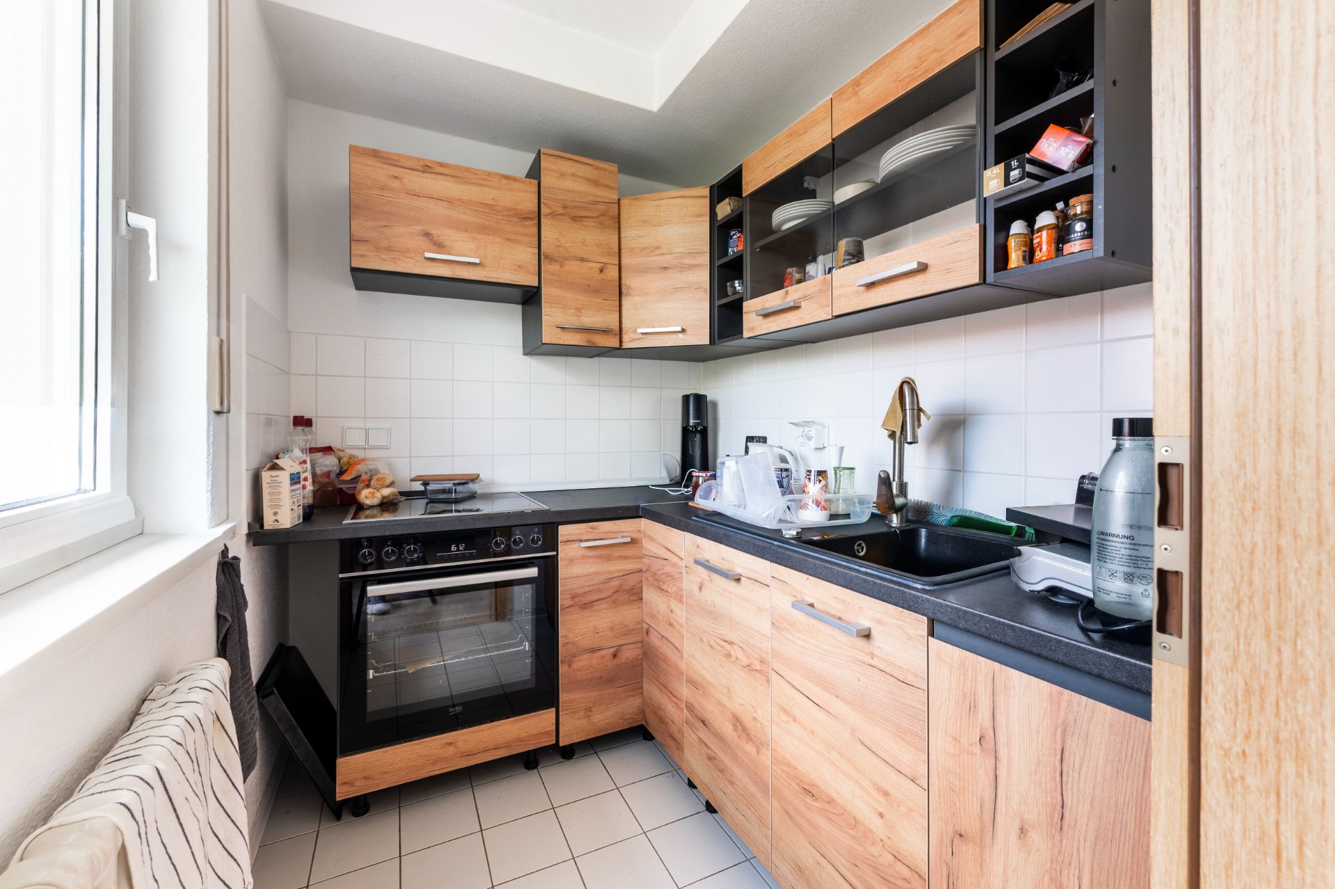 Moderne Einbauküche mit Holzfronten, Elektroherd, Spüle und offenem Regal, Tageslicht durch Fenster.