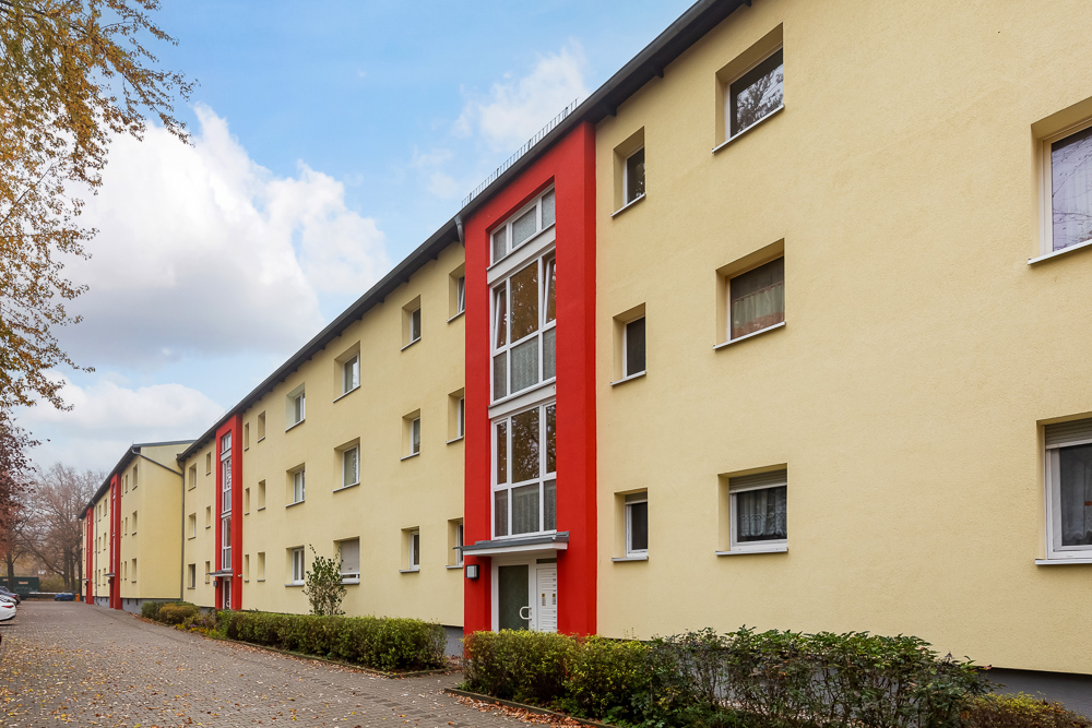 Mehrfamilienhaus mit gelber Fassade und roten Akzenten, viele Fenster und Sträucher davor.