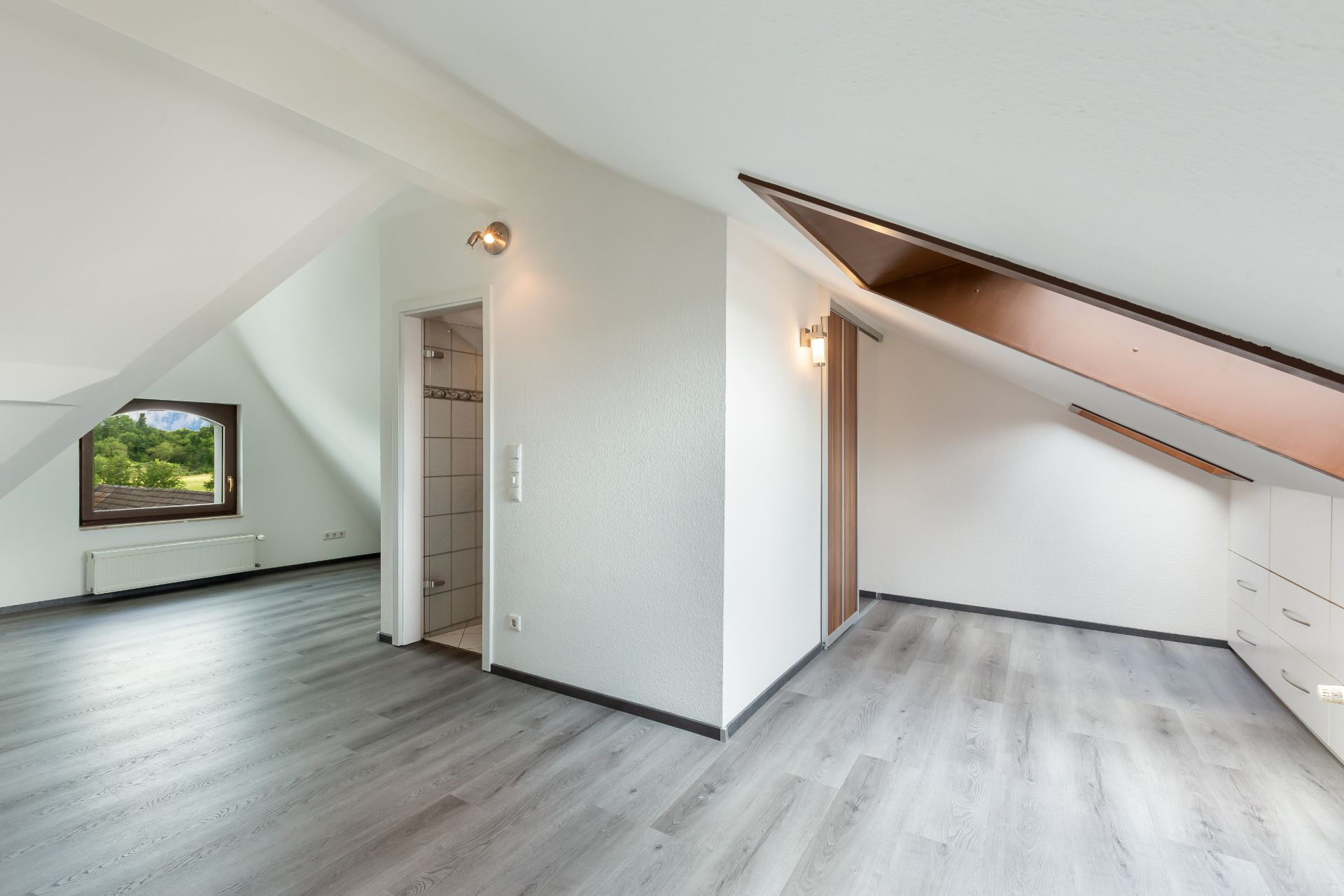 Helles Dachzimmer mit großem Fenster, Holzfußboden und Zugang zu einem kleinen Badezimmer.