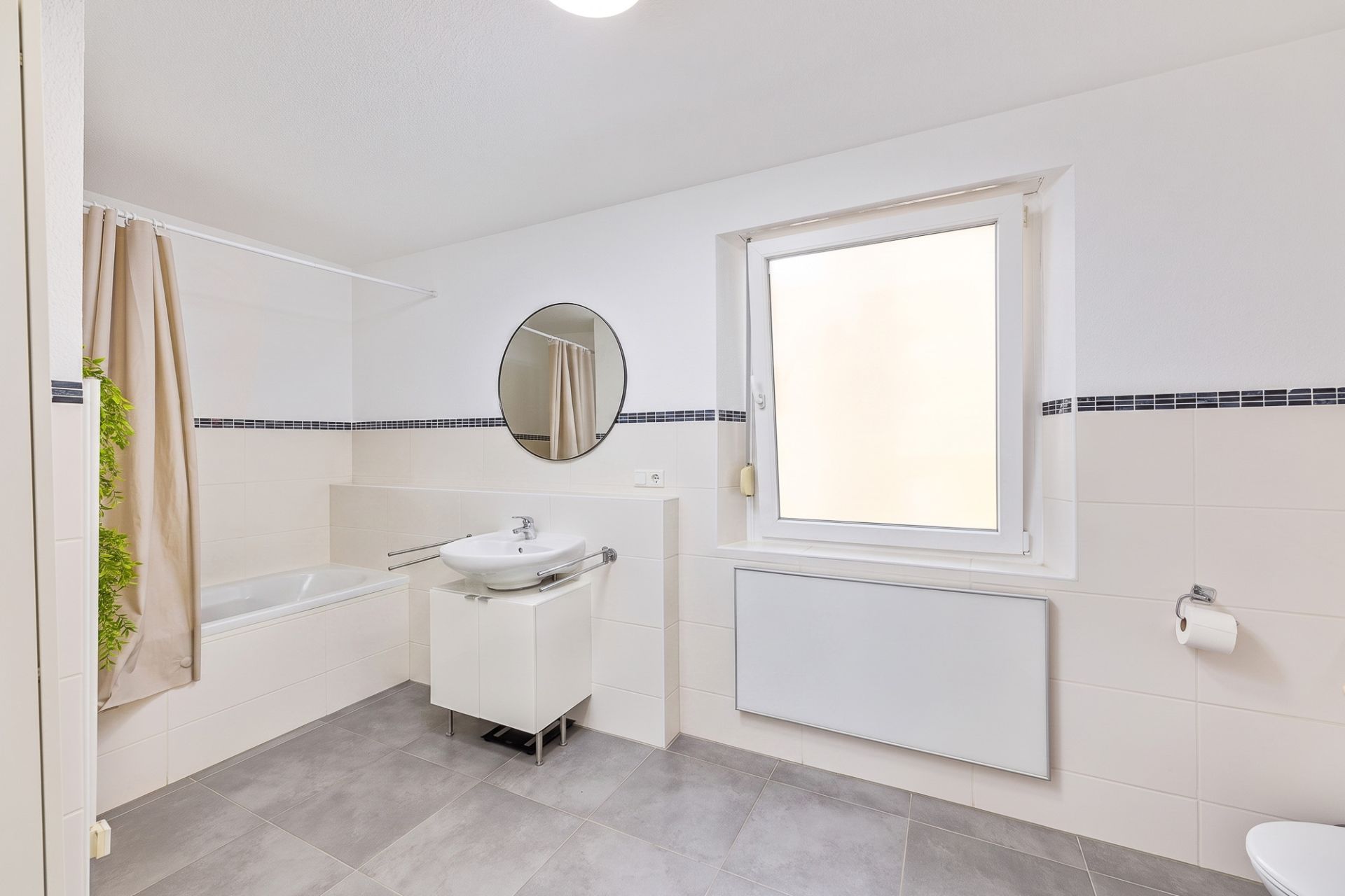 Helles Badezimmer mit Badewanne, Waschbecken, rundem Spiegel und Fenster.