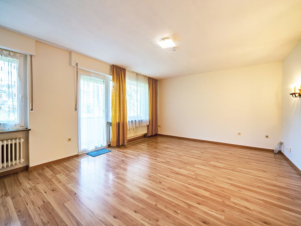 Leeres Wohnzimmer mit Holzboden, großen Fenstern und gelben Vorhängen.