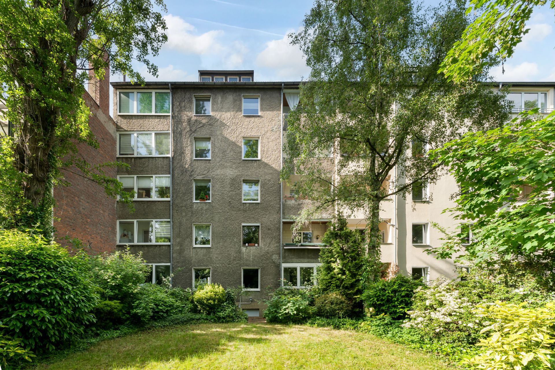 Mehrstöckiges Wohnhaus mit mehreren Fenstern, umgeben von Bäumen und Grünfläche.