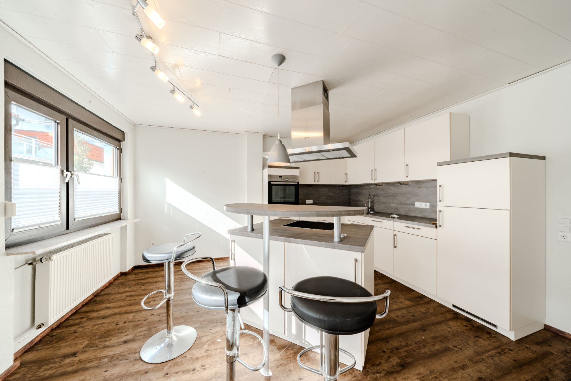 Helle moderne Küche mit Tresen, drei Barhockern, Einbauschränken und Fenstern, Holzboden.