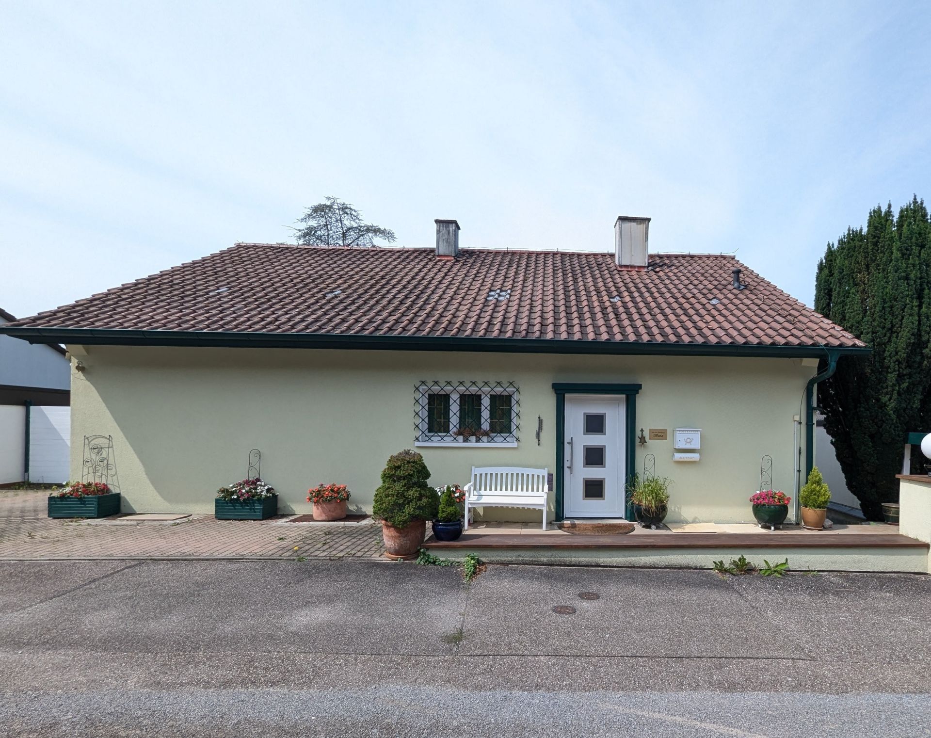 Vorderansicht eines Hauses mit rotem Dach, Bank und Blumenkübeln vor dem Eingang.