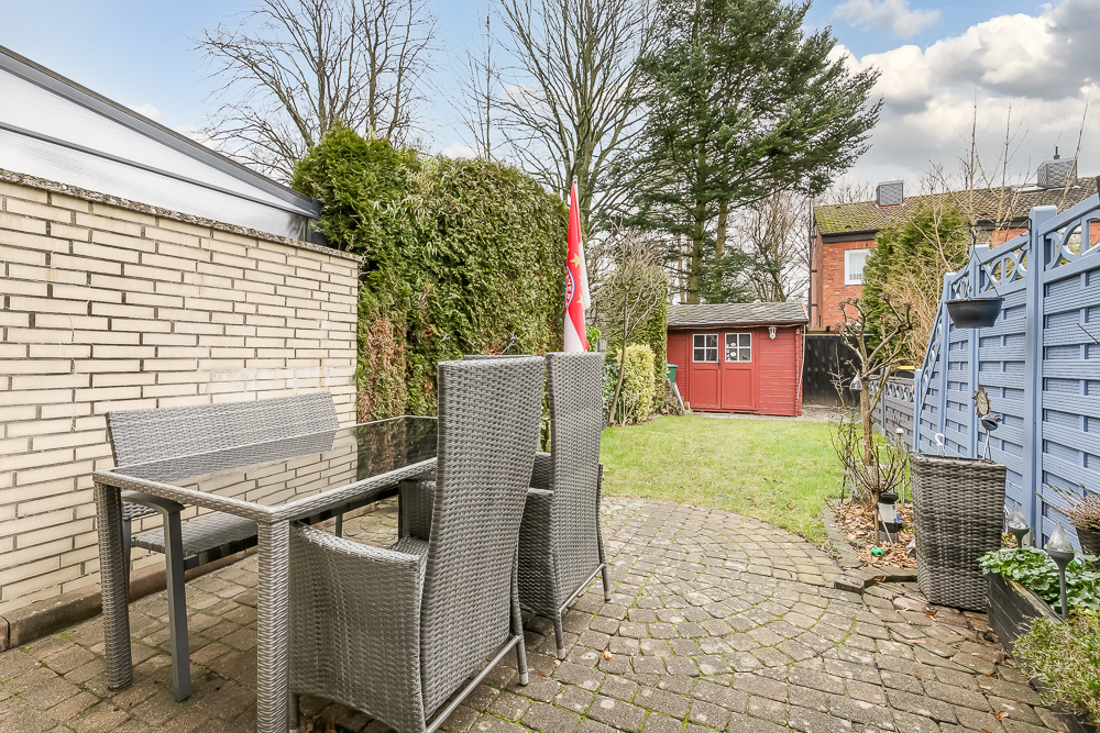 Garten mit Terrasse, Gartentisch, Stühlen und einem roten Gartenhaus im Hintergrund.