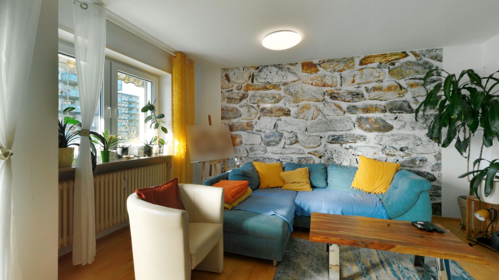Helles Wohnzimmer mit Ecksofa, Sesseln, Holztisch und Pflanze vor einer Steinwand.
