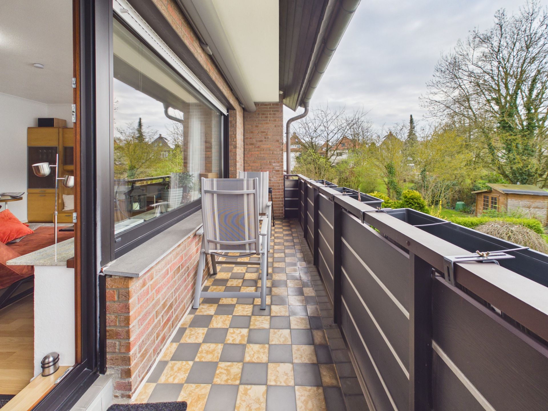 Langer Balkon mit gefliesten Boden, Stuhl und Blick auf Garten und Bäume.
