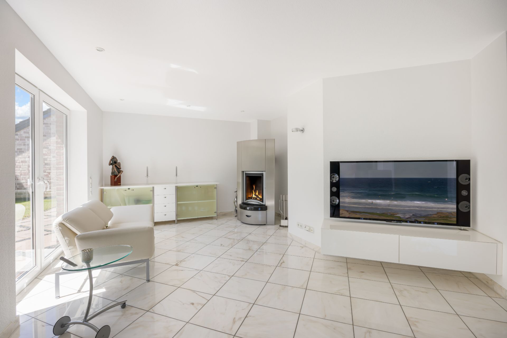 Helles Wohnzimmer mit modernem Kamin, Flachbildfernseher, Sofa und Glastisch auf Fliesenboden.