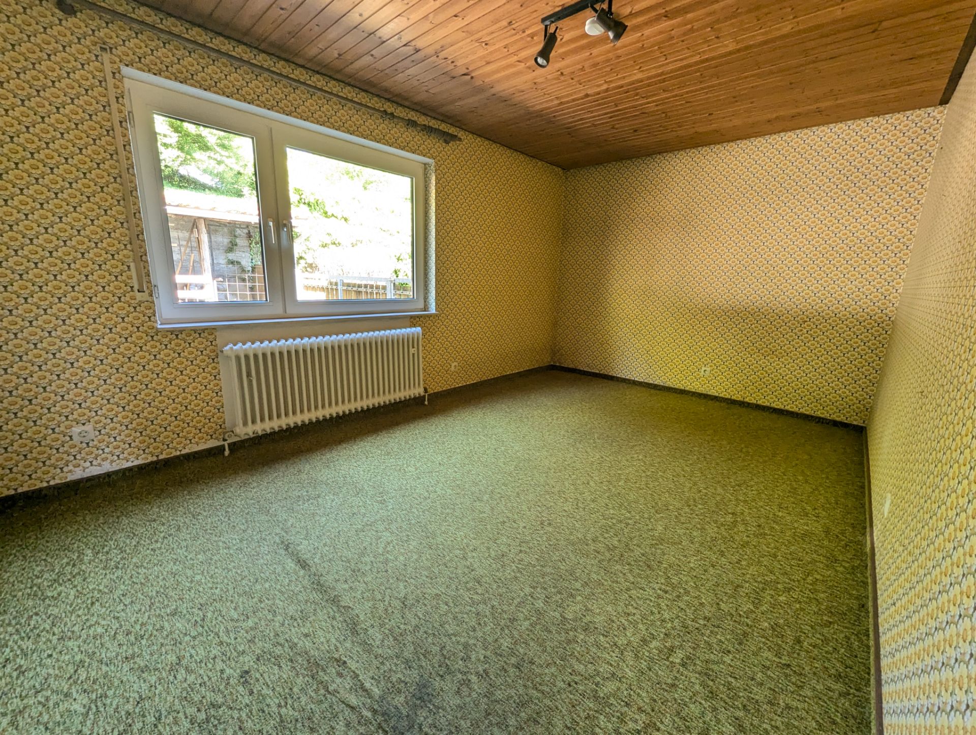 Leerer Raum mit gemusterten Wänden, Teppichboden, Holzdecke und großem Fenster mit Heizung.