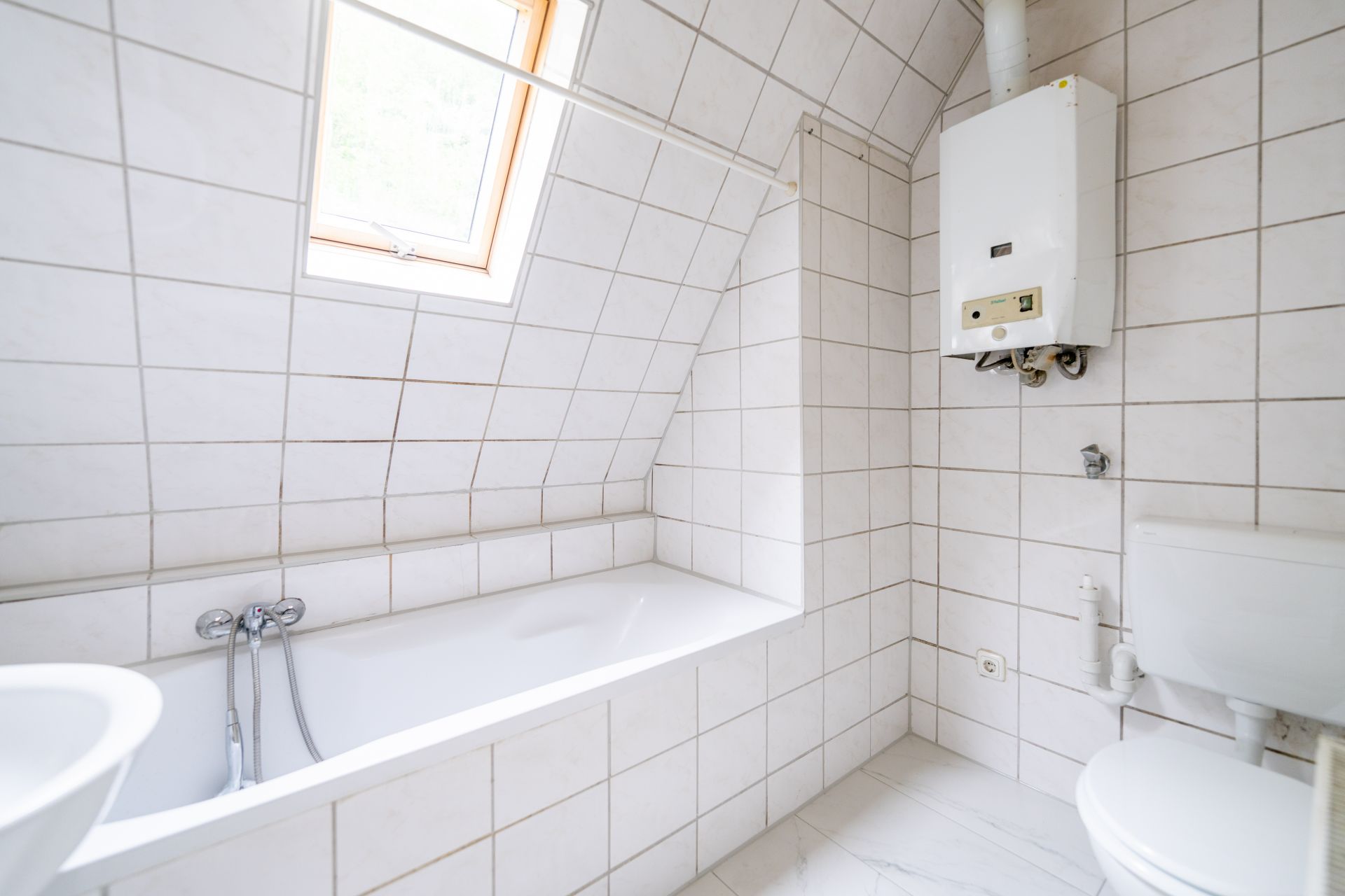 Helles Badezimmer mit Badewanne, Fenster, weiß gefliesten Wänden und einem Boiler an der Wand.