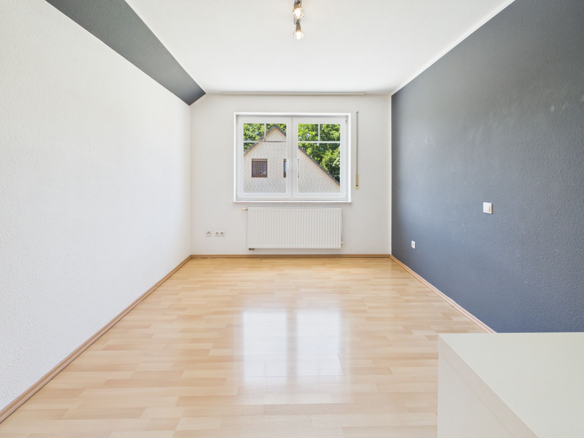 Leerer Raum mit hellem Laminatboden, weißen Wänden und einem Fenster an der Stirnseite.