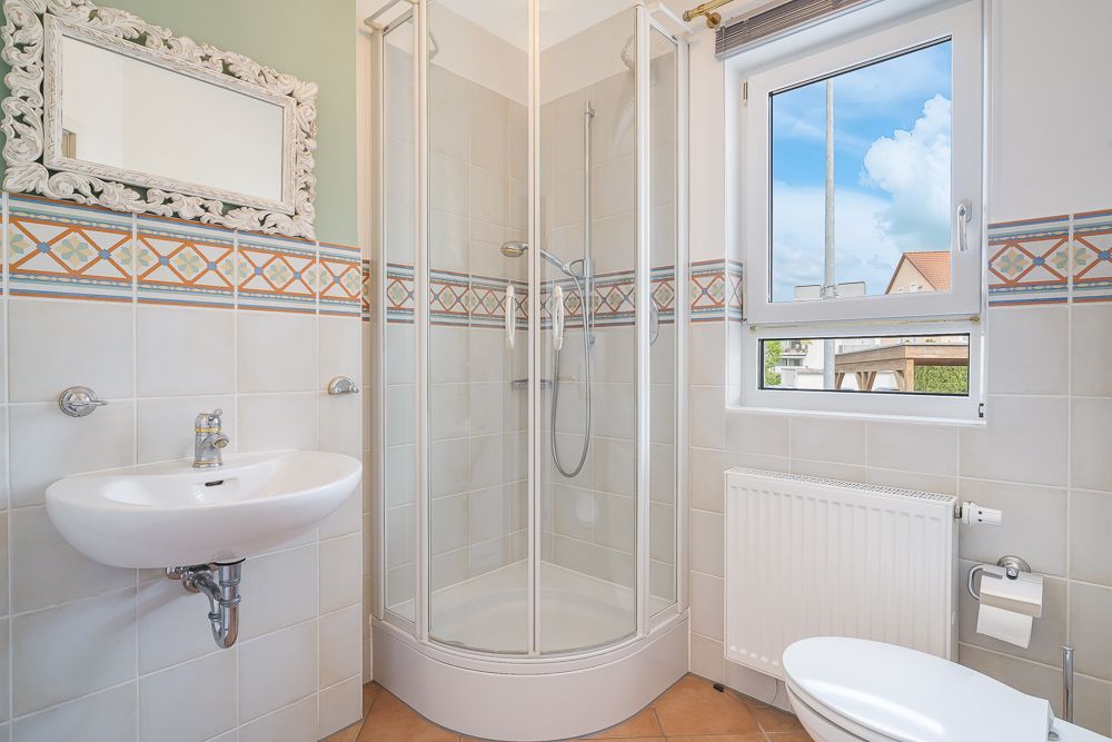 Helles Badezimmer mit Dusche, Waschbecken, Spiegel, Fenster, Heizung und Toilette.