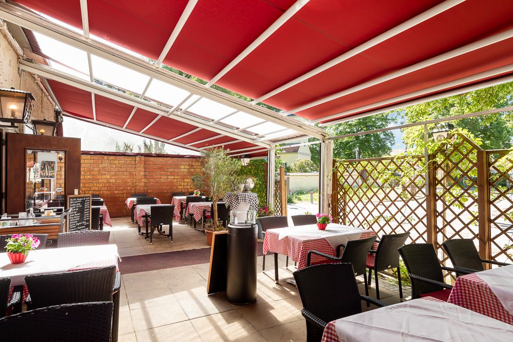 Überdachte Terrasse mit Tischen, Stühlen und rot-weiß karierten Tischdecken im Garten.