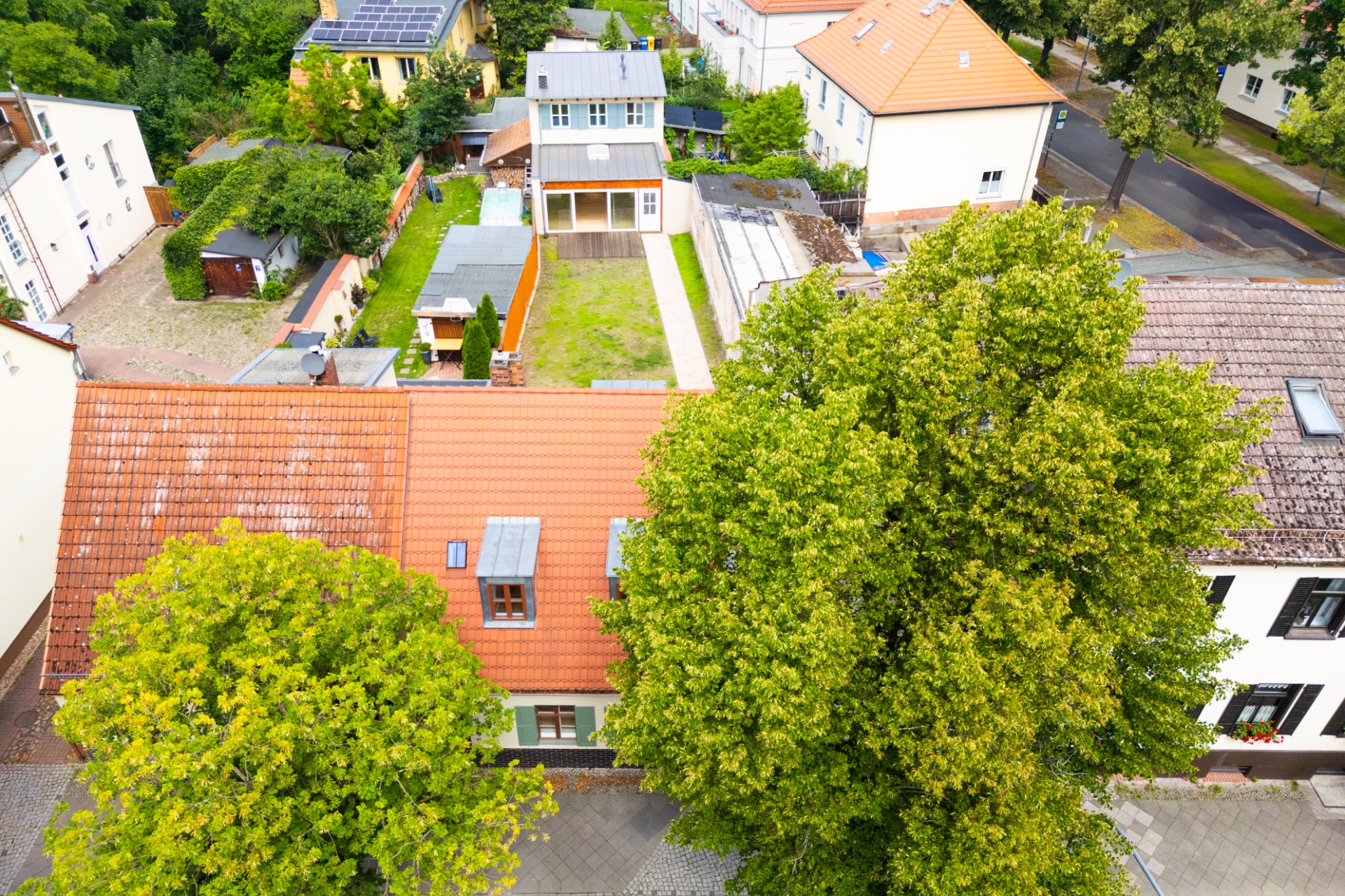 Luftaufnahme eines Wohnhauses mit Garten und mehreren Nebengebäuden, Bäume im Vordergrund.