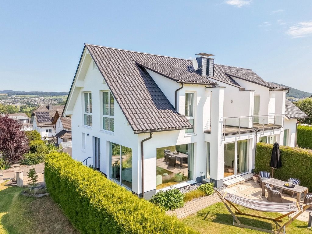 Modernes weißes Einfamilienhaus mit Garten, Terrasse und großen Fenstern an einem sonnigen Tag.
